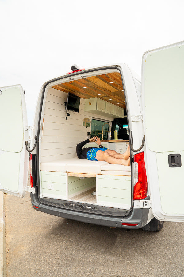 Open back of an RV rental: a person relaxes inside a custom camper van with a cozy bed, TV, and wood-paneled ceiling.