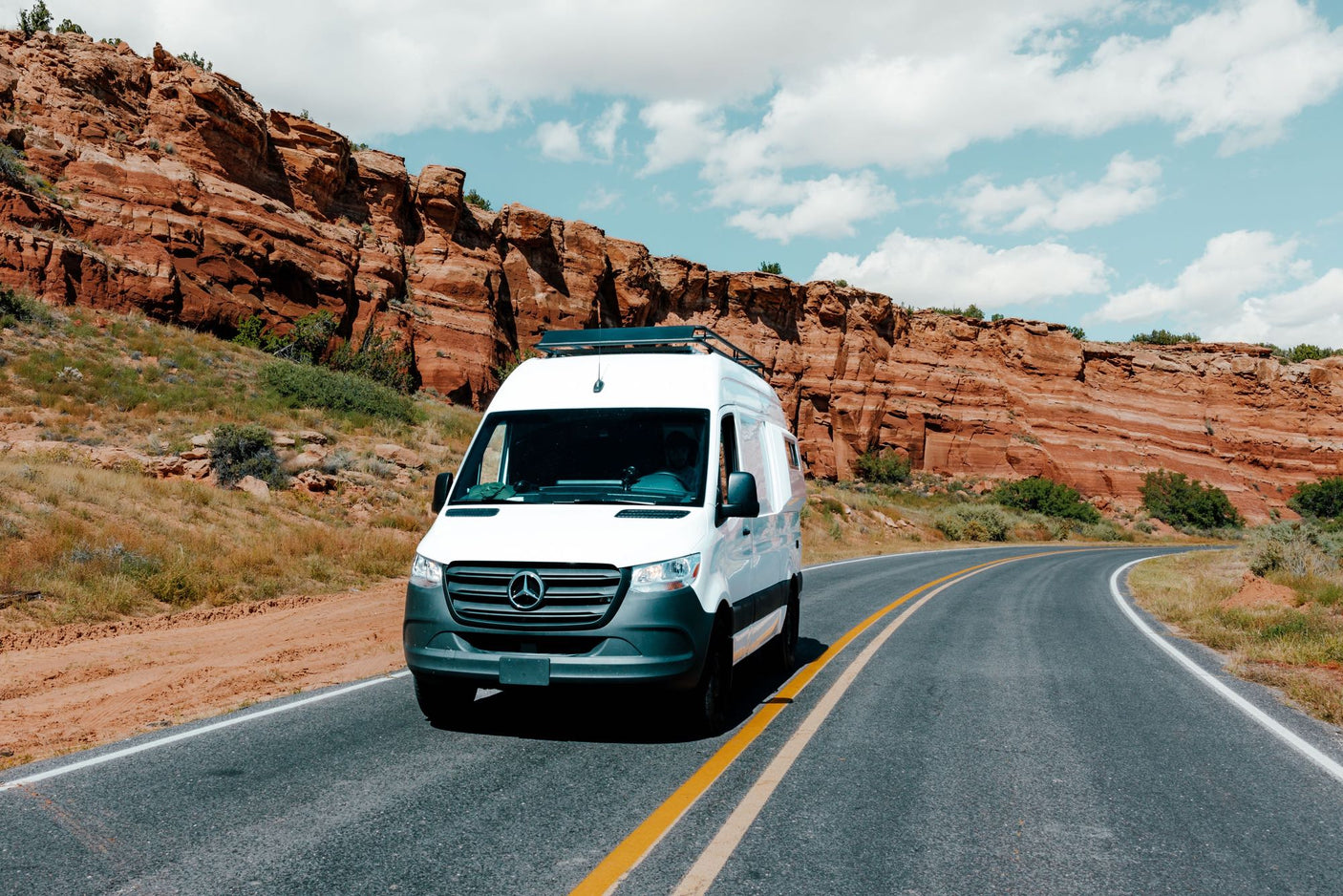 A white Mercedes-Benz Sprinter van, perfect for an rv rental adventure, drives past towering red rock cliffs on a scenic road.