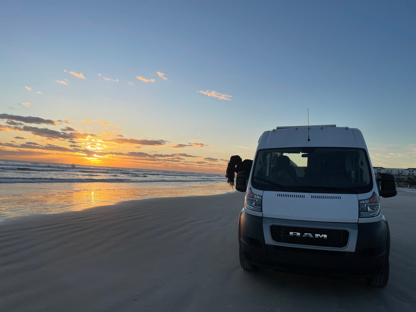 White rv rental van on a beach at sunset. Person leans out to see golden ocean & sky views.