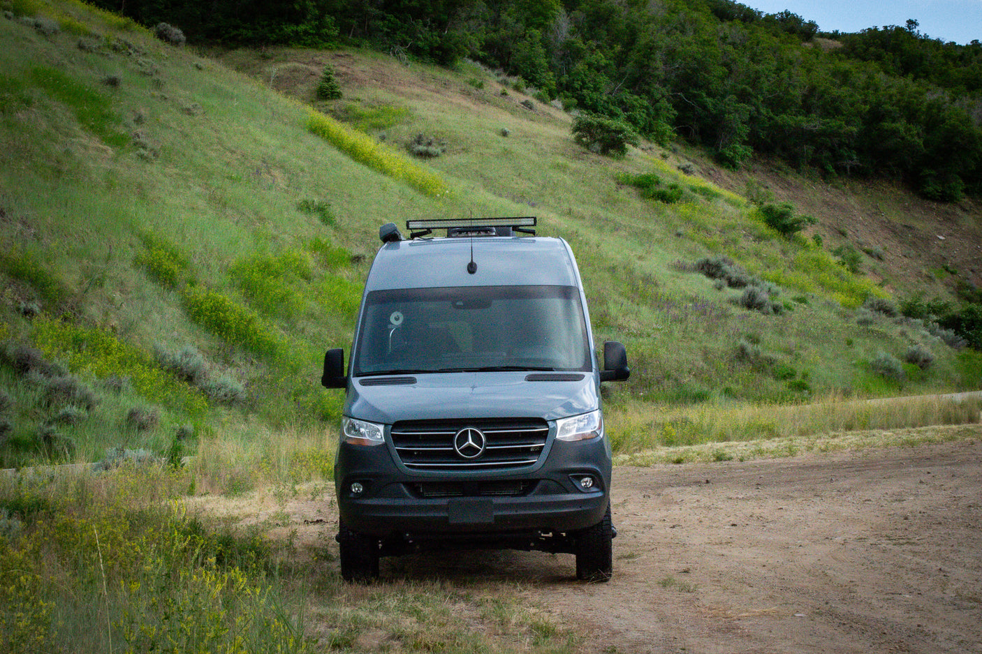 Gray Mercedes-Benz camper van parked on a dirt road with a lush green hill and trees in the background. Perfect for an RV rental adventure.