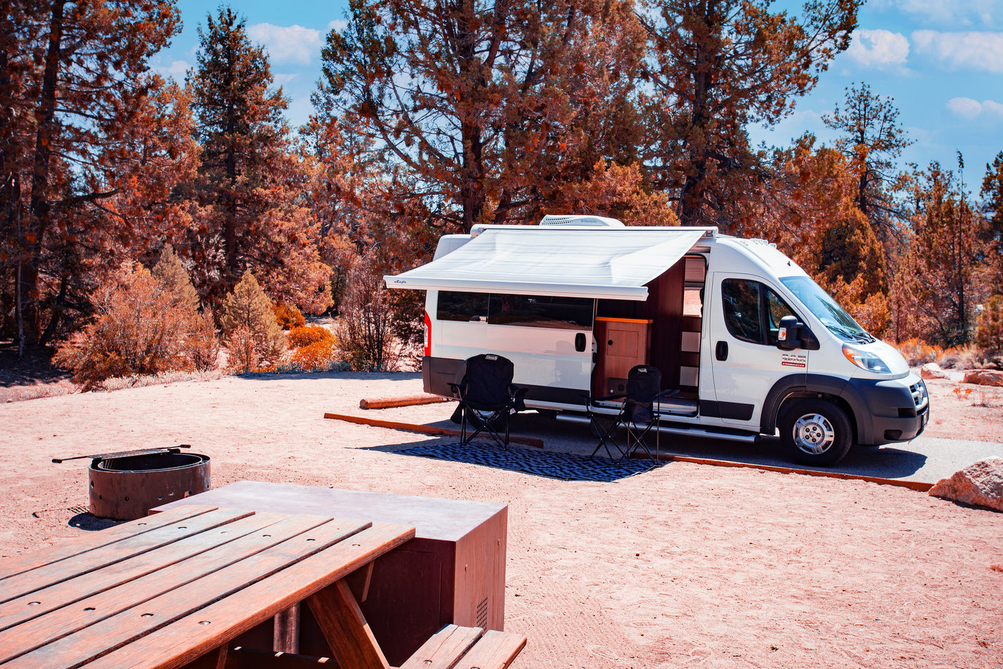 RV rental campervan parked at a scenic campsite with an extended awning, chairs, picnic table, and fire pit setup.