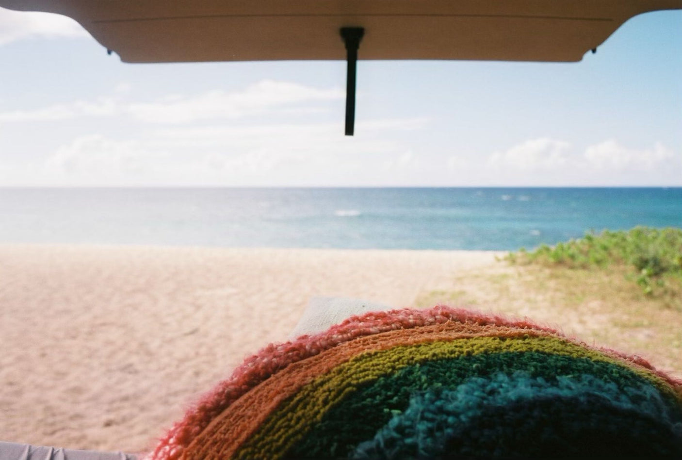 Hatchback view from an **RV rental**: serene beach, turquoise ocean, & a cozy rainbow blanket.