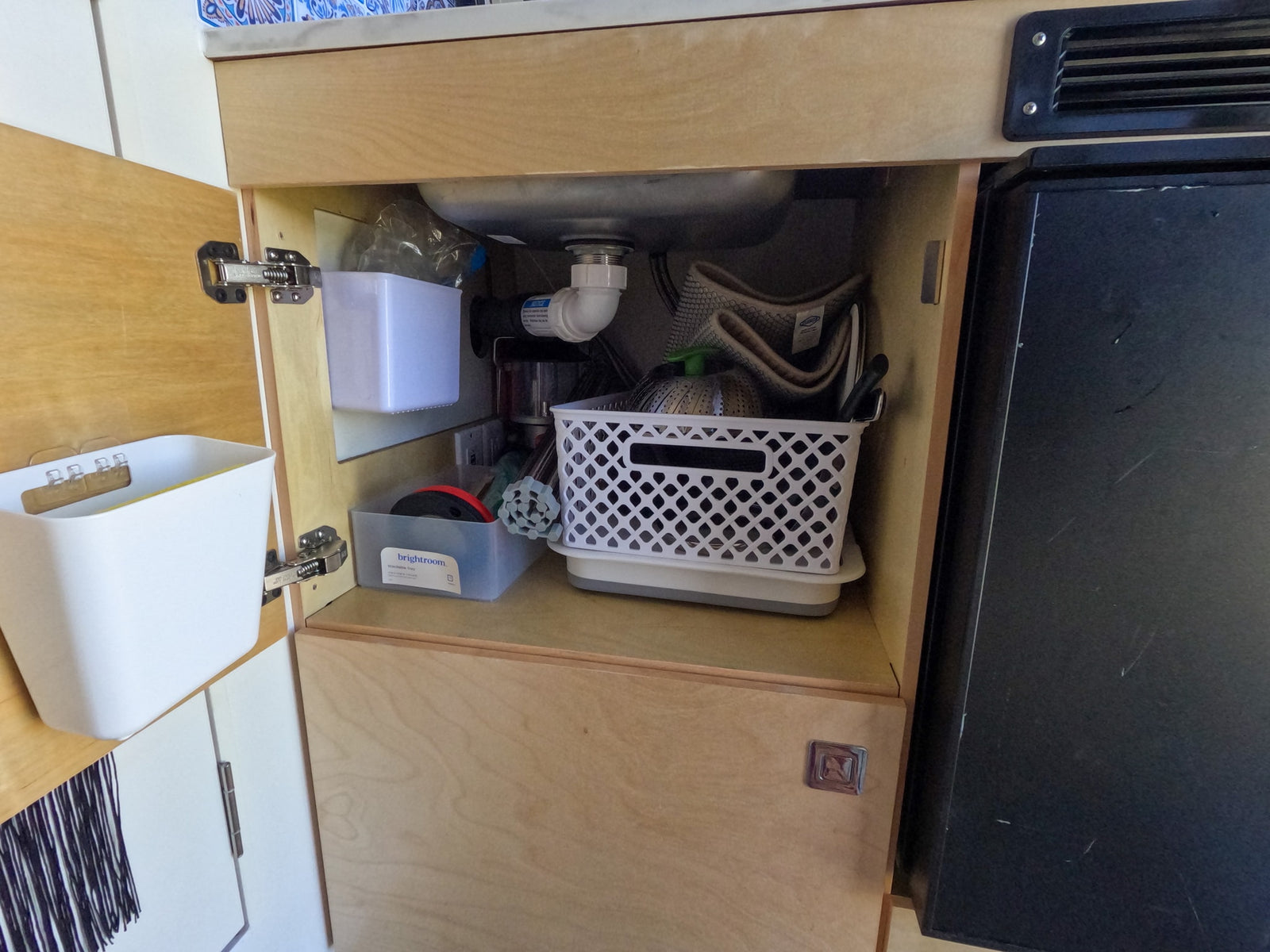 Organized under-sink cabinet in an rv rental kitchen with storage bins, water filter, and sink plumbing next to a fridge.