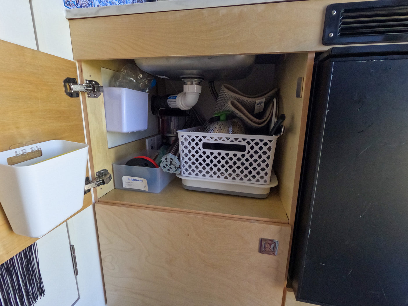 Organized under-sink cabinet in an rv rental kitchen with storage bins, water filter, and sink plumbing next to a fridge.