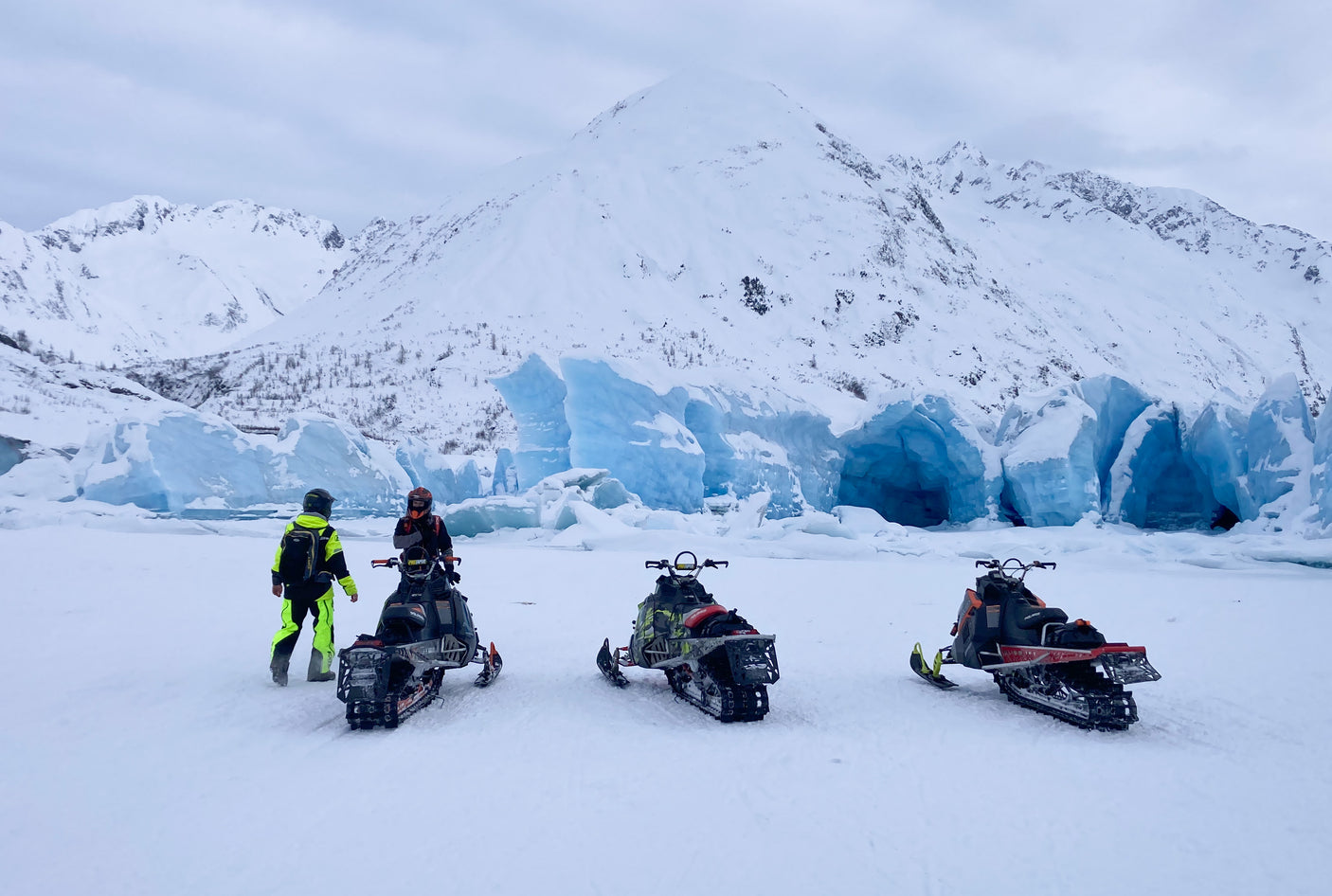 Three snowmobiles and two adventurers face a vibrant blue glacier and snowy mountains. Discover winter with an RV rental!
