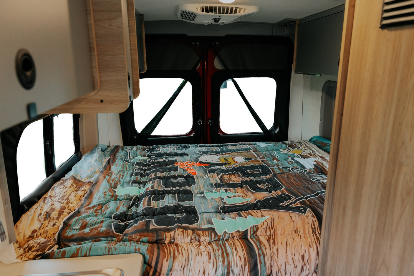 RV rental interior: a comfortable bed with a colorful patterned comforter, visible bright windows at the back.