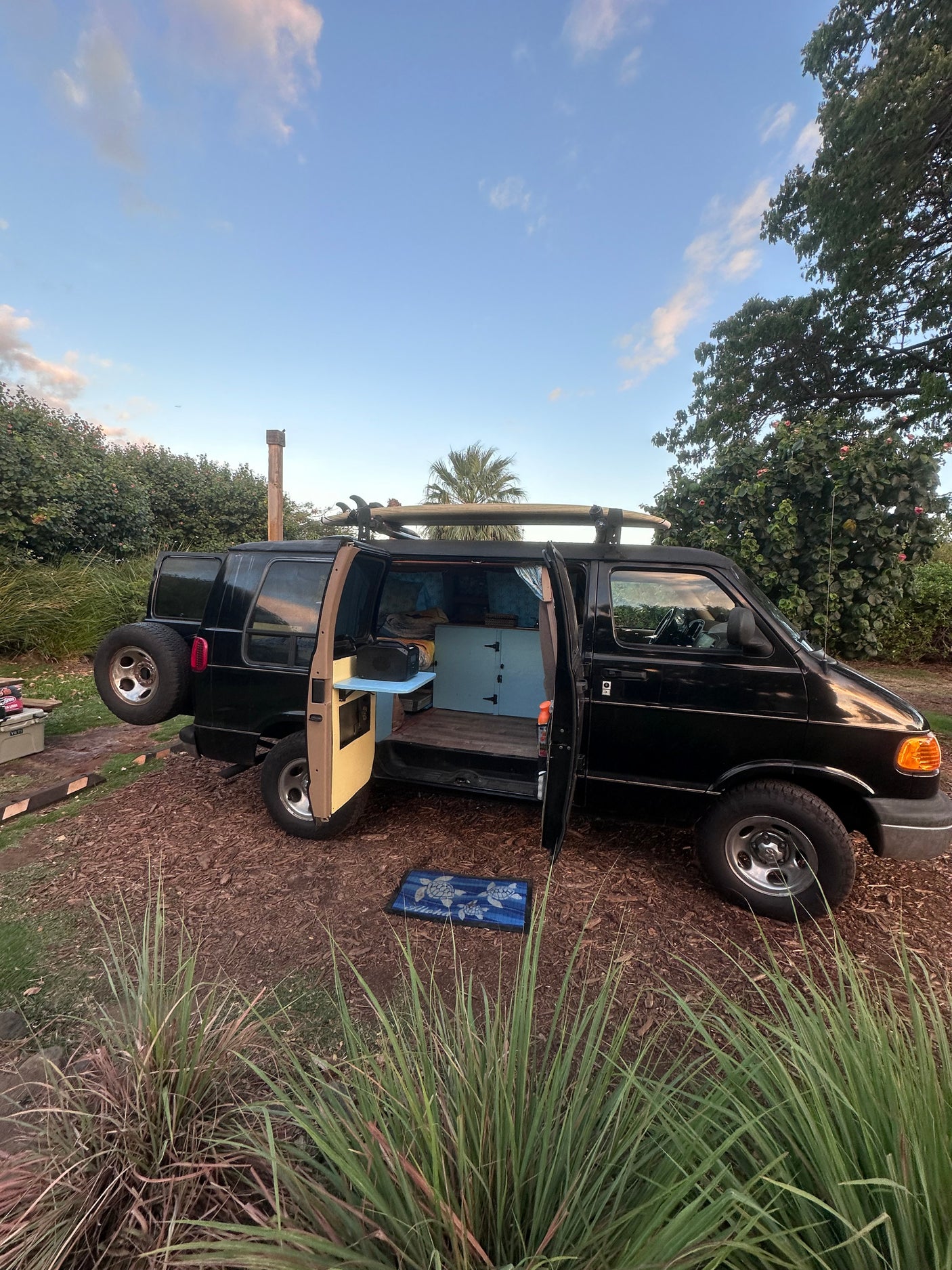 This black camper van, an RV rental, with surfboards on top, doors open revealing its custom interior. Parked for adventure.