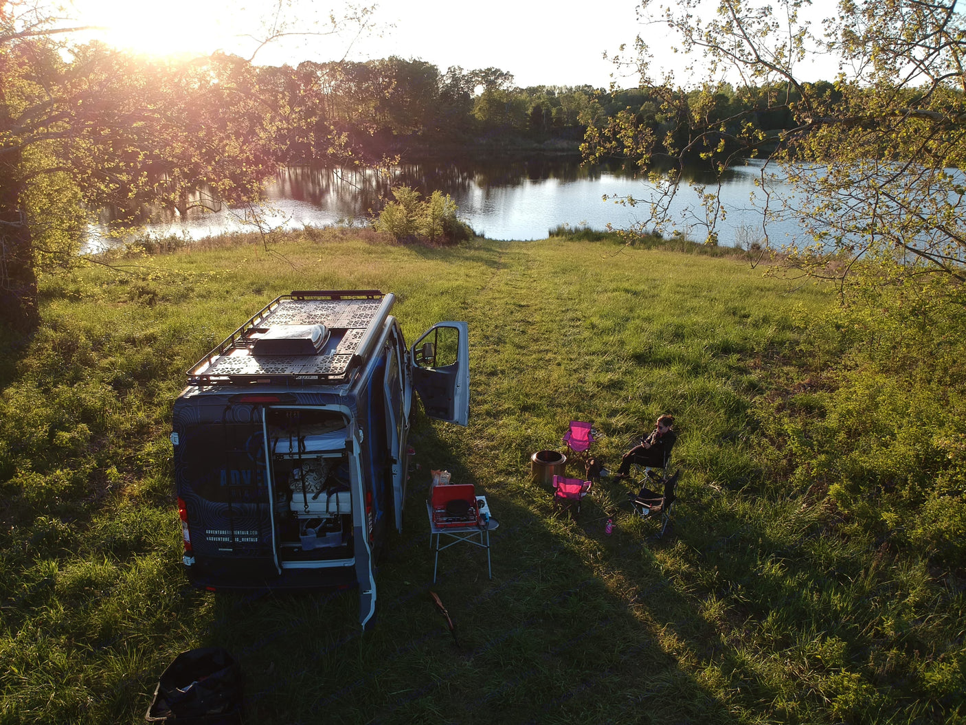 Overhead view: an RV rental van is parked on grassy hill beside a lake, with a person by a fire pit & chairs at sunset.