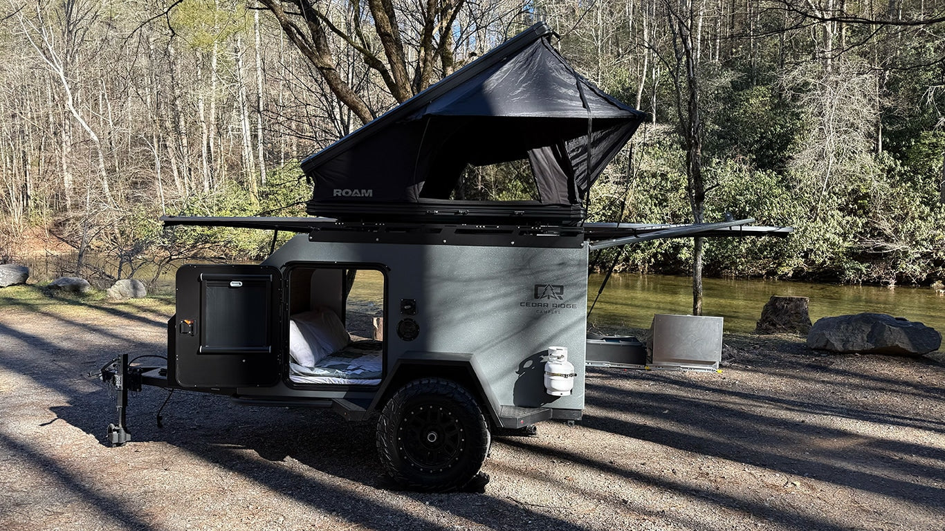 Compact Cedar Ridge Campers rv rental with a deployed ROAM rooftop tent by a serene river, perfect for outdoor adventure.