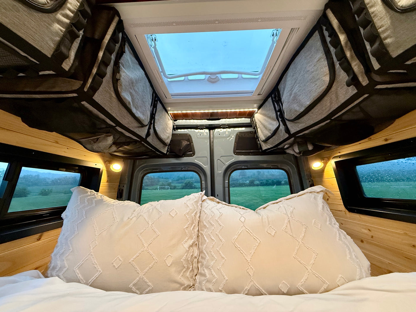 Cozy RV rental interior: bed with pillows, wood walls, skylight, and scenic view from the back.