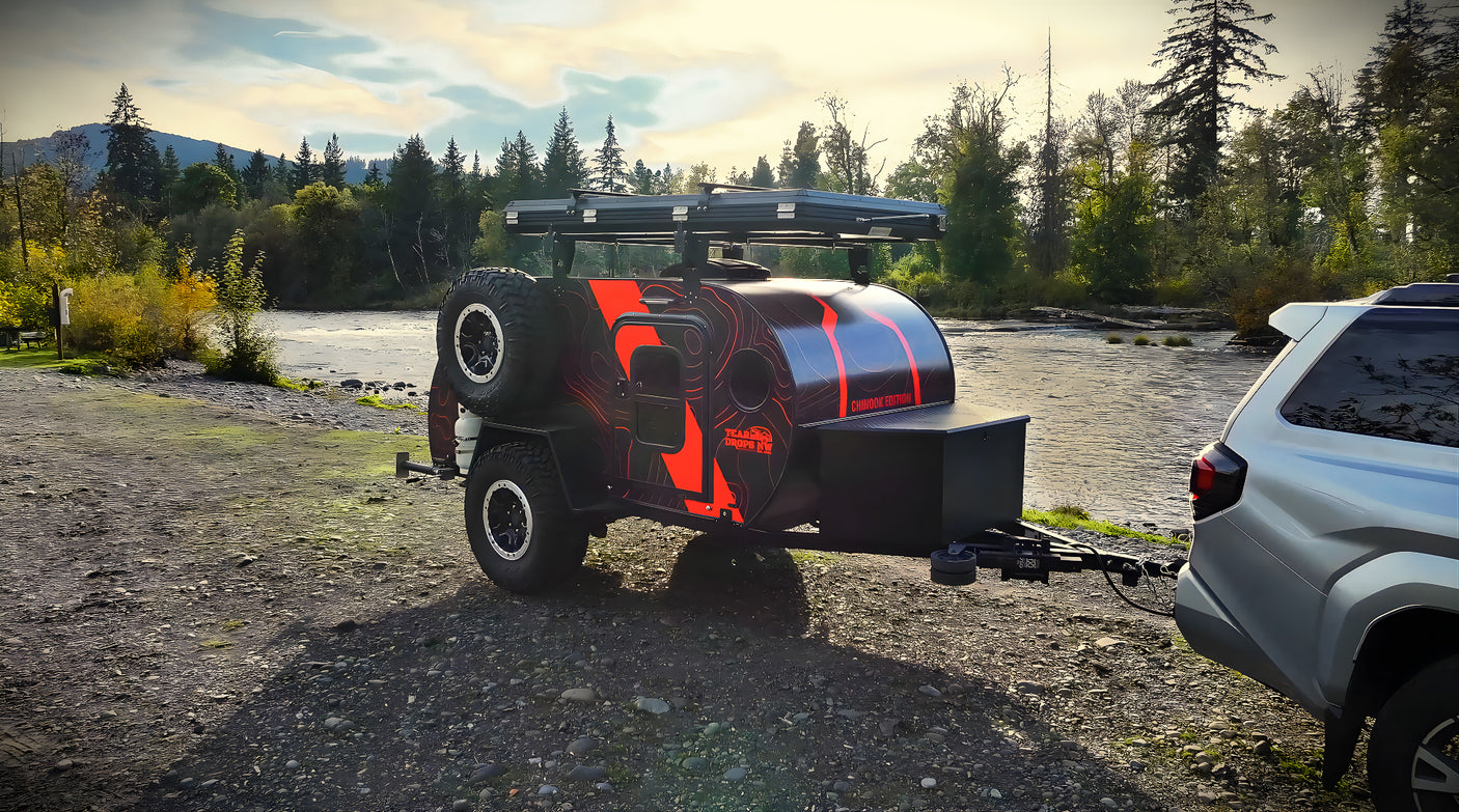 An off-road teardrop trailer, ideal for an RV rental, parked by a river in a scenic mountain setting.