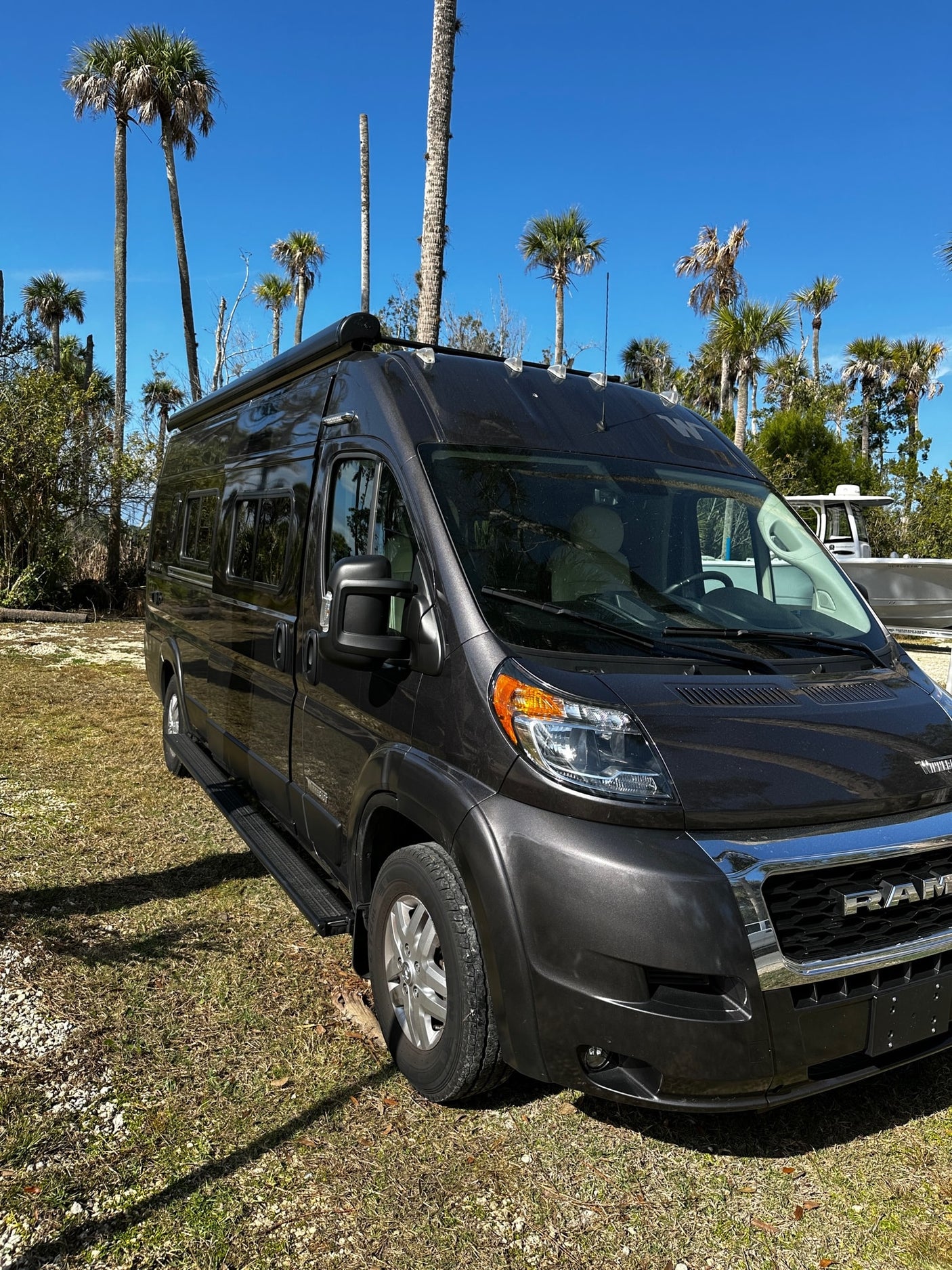 Dark grey Ram ProMaster camper van, ideal for an RV rental, parked on grass near palm trees under a blue sky.