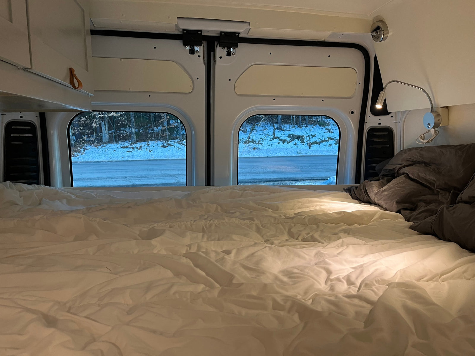 Cozy rv rental interior: A bed with white sheets and gray bedding, offering a view of a snowy road and forest for winter travel.