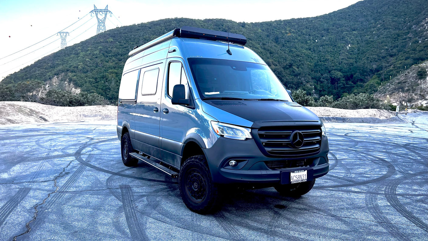 This rugged blue Mercedes-Benz Sprinter camper van, an ideal rv rental, is ready for adventure, parked before a forested mountain.