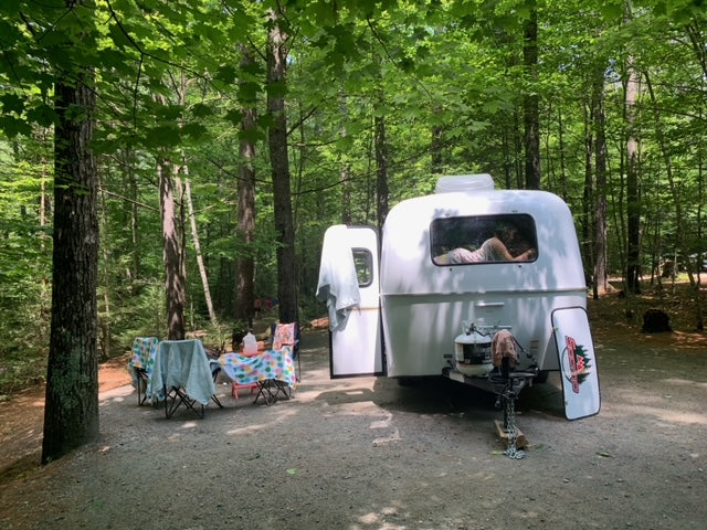 A person relaxes inside a white teardrop RV rental parked in a shaded forest campsite with a picnic area.