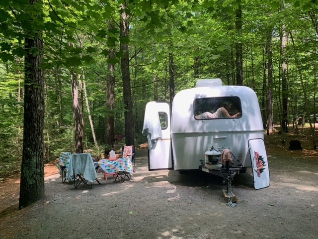 parked in grass. A great RV rental option.|A person relaxes inside a white teardrop RV rental parked in a shaded forest campsite with a picnic area.|Woman enjoys nature view from RV rental window. Cozy camper interior features kitchen