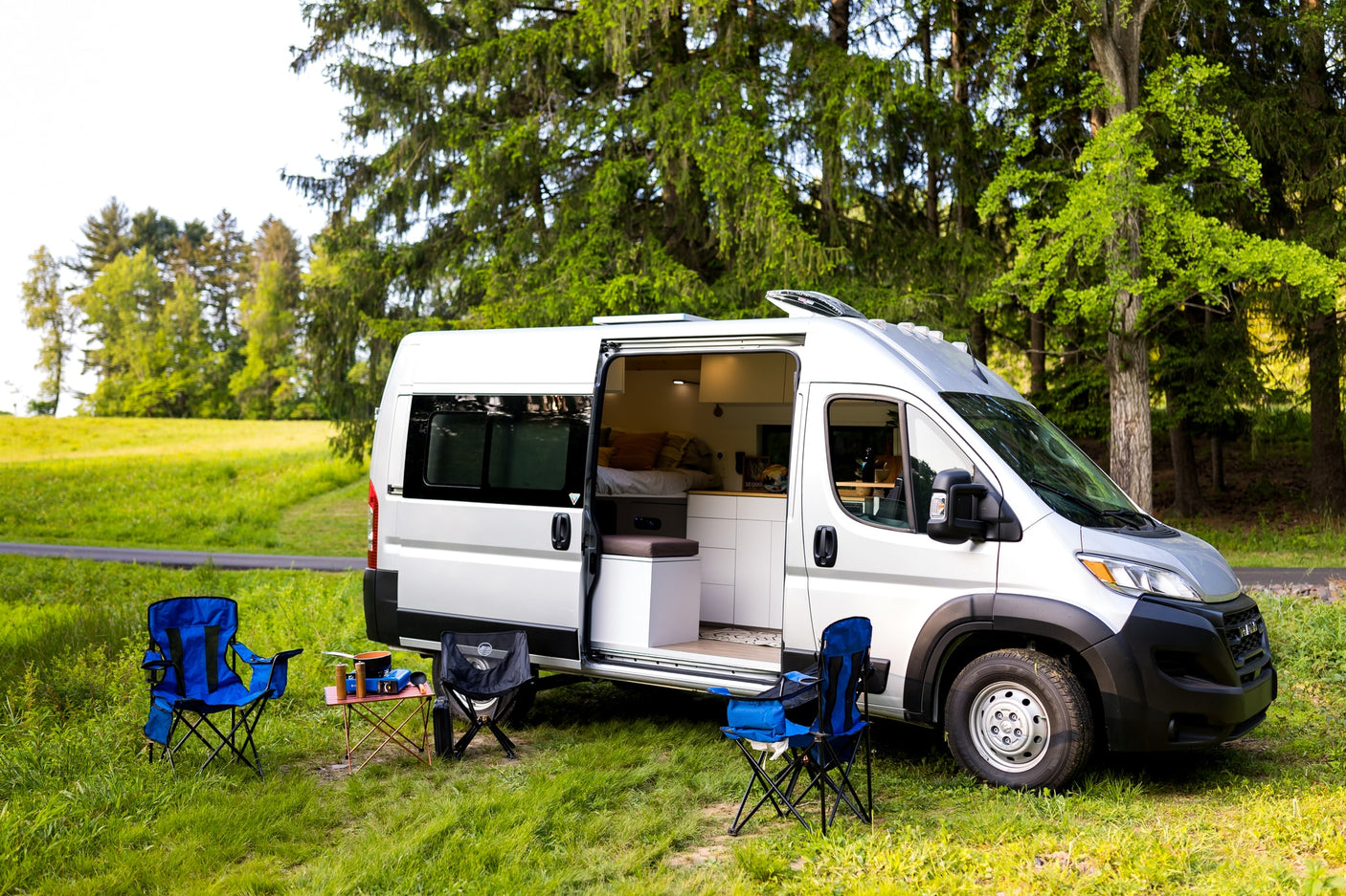 Silver camper van RV rental parked in grass, side door open. Camping chairs & cook set up outside for an outdoor adventure.
