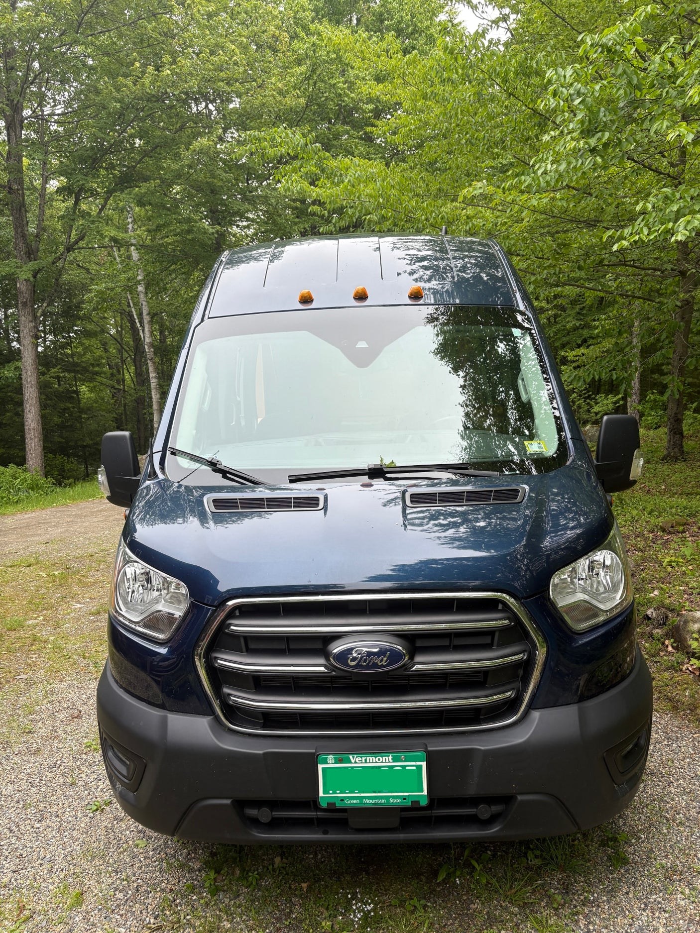 This blue Ford Transit van RV rental is parked on a gravel path, surrounded by lush green trees.