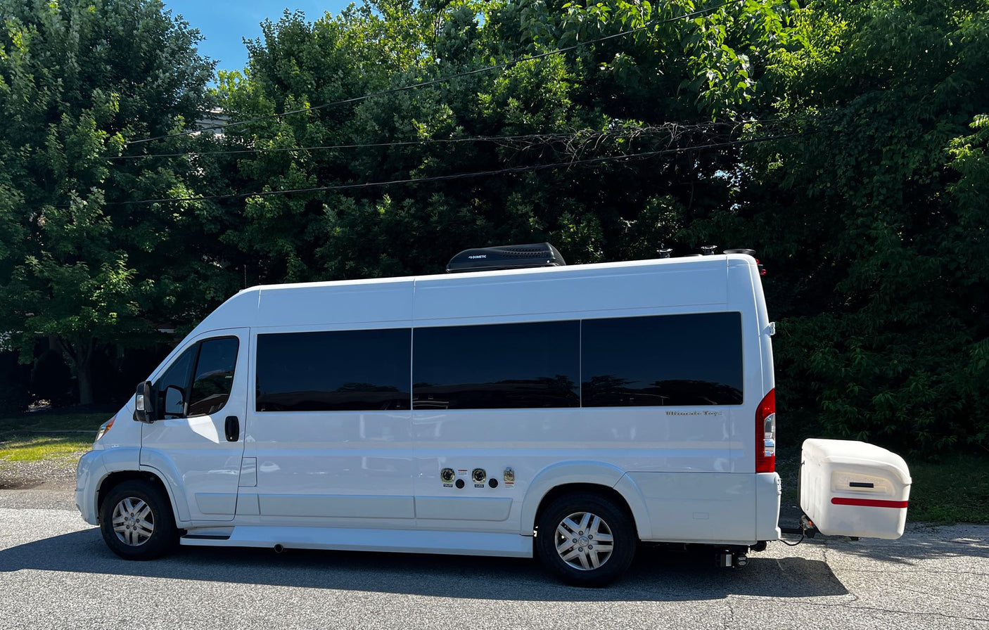 White RV rental camper van parked roadside. Features tinted windows, a Dometic roof unit, and a rear hitch-mounted storage box.