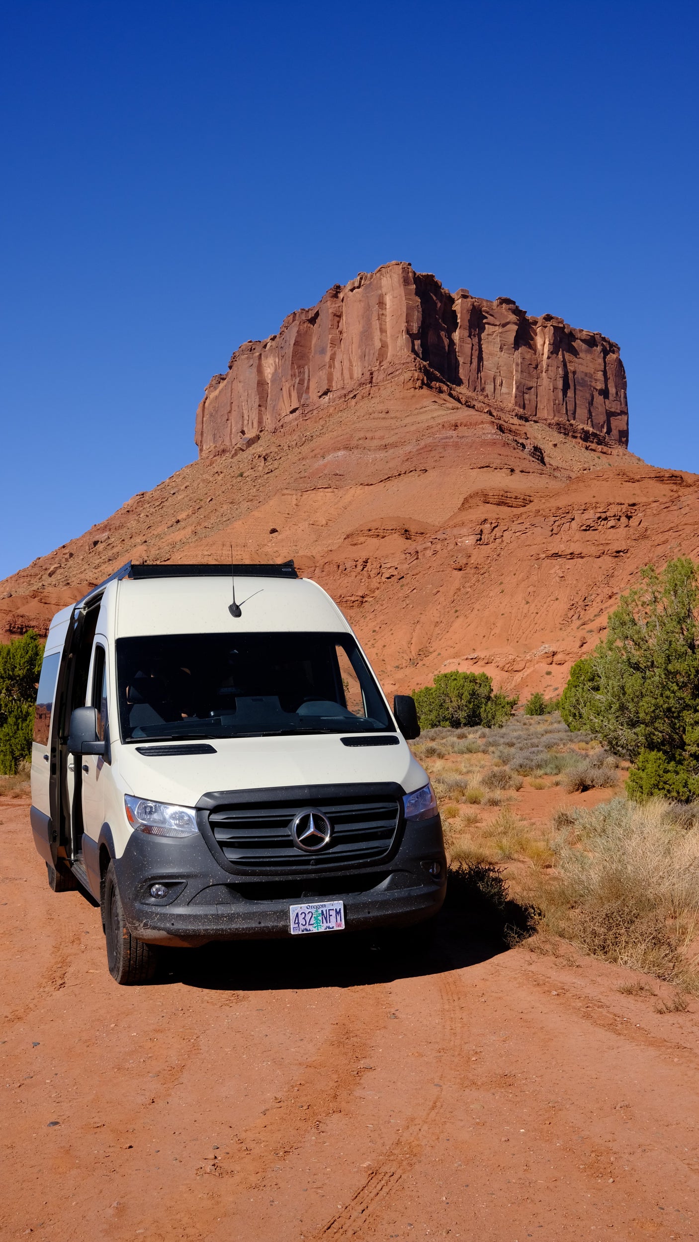 Scenic RV rental adventure: A white Mercedes Sprinter campervan on a red dirt road with massive red rock formations under a blue sky.