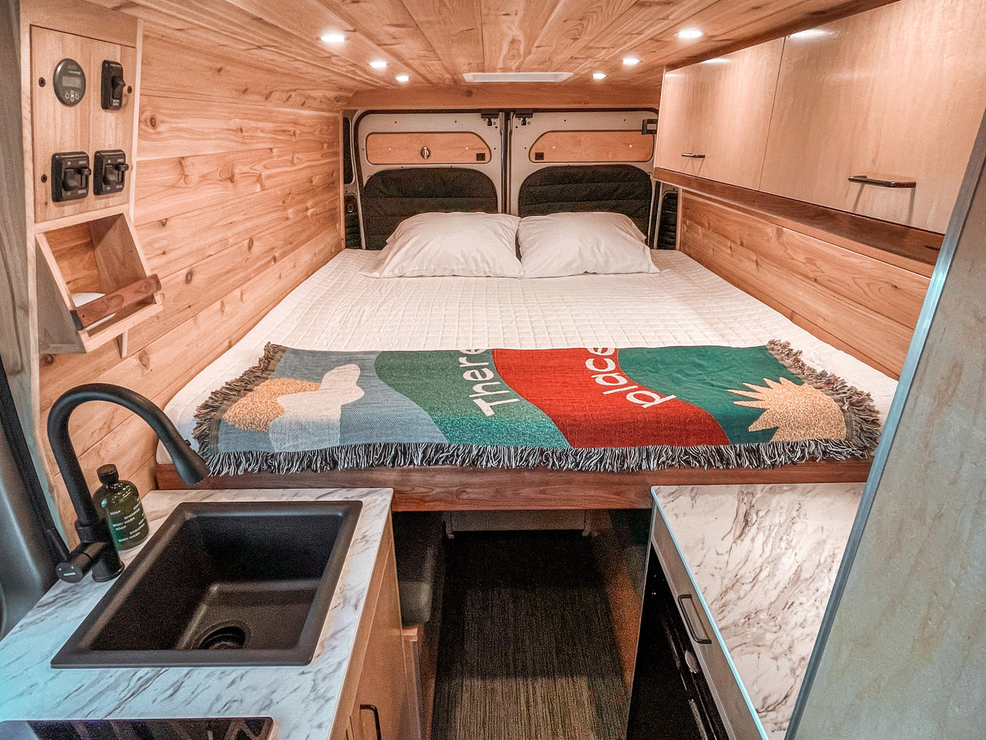 Cozy RV rental interior with a comfortable bed, custom wood paneling, overhead storage, and a functional kitchen sink area.