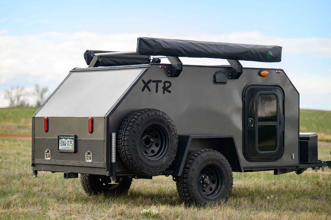A rugged dark gray XTR off-road camper trailer in a grassy field. An ideal RV rental for backcountry adventures.