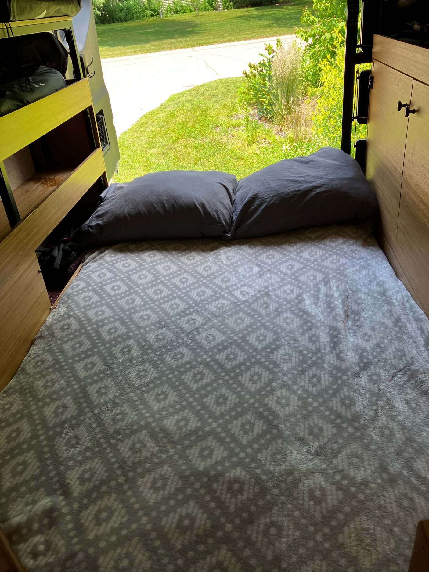 Comfortable RV rental bed with grey pillows & patterned blanket, looking out to a sunny green yard. Ideal for road trip rest.