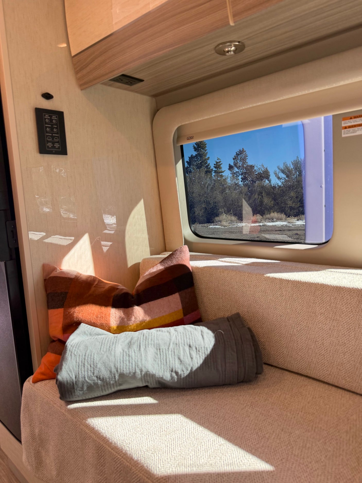 Cozy RV rental interior with a comfy bench, patterned pillows, and a scenic window view of trees and blue sky.