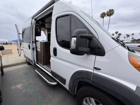 White RV rental parked at the beach with its side door open, revealing the interior kitchenette. Palm trees dot the cloudy horizon.
