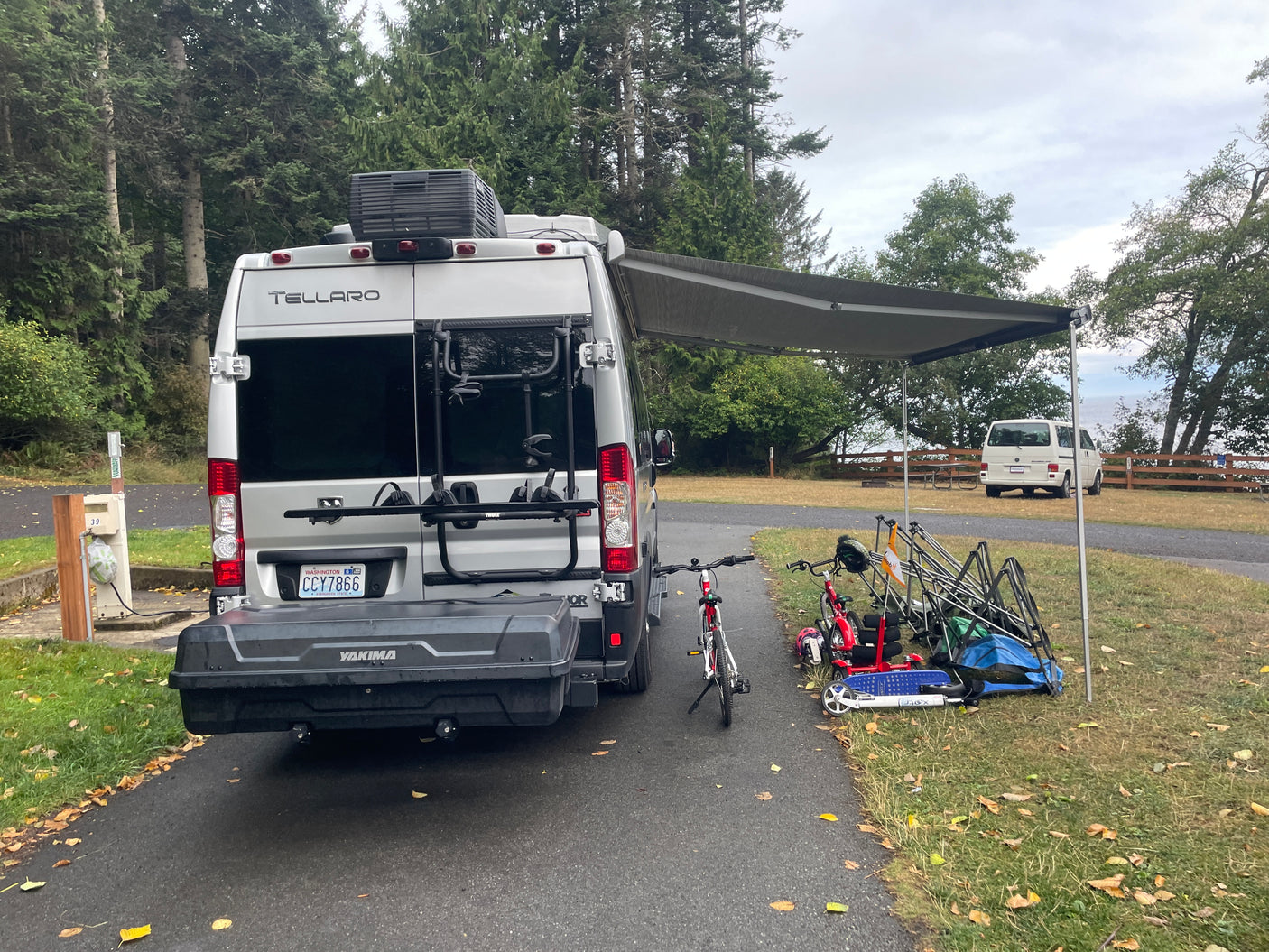 RV rental (Tellaro) parked at a campsite, awning extended, with bikes, scooters, and chairs outside.