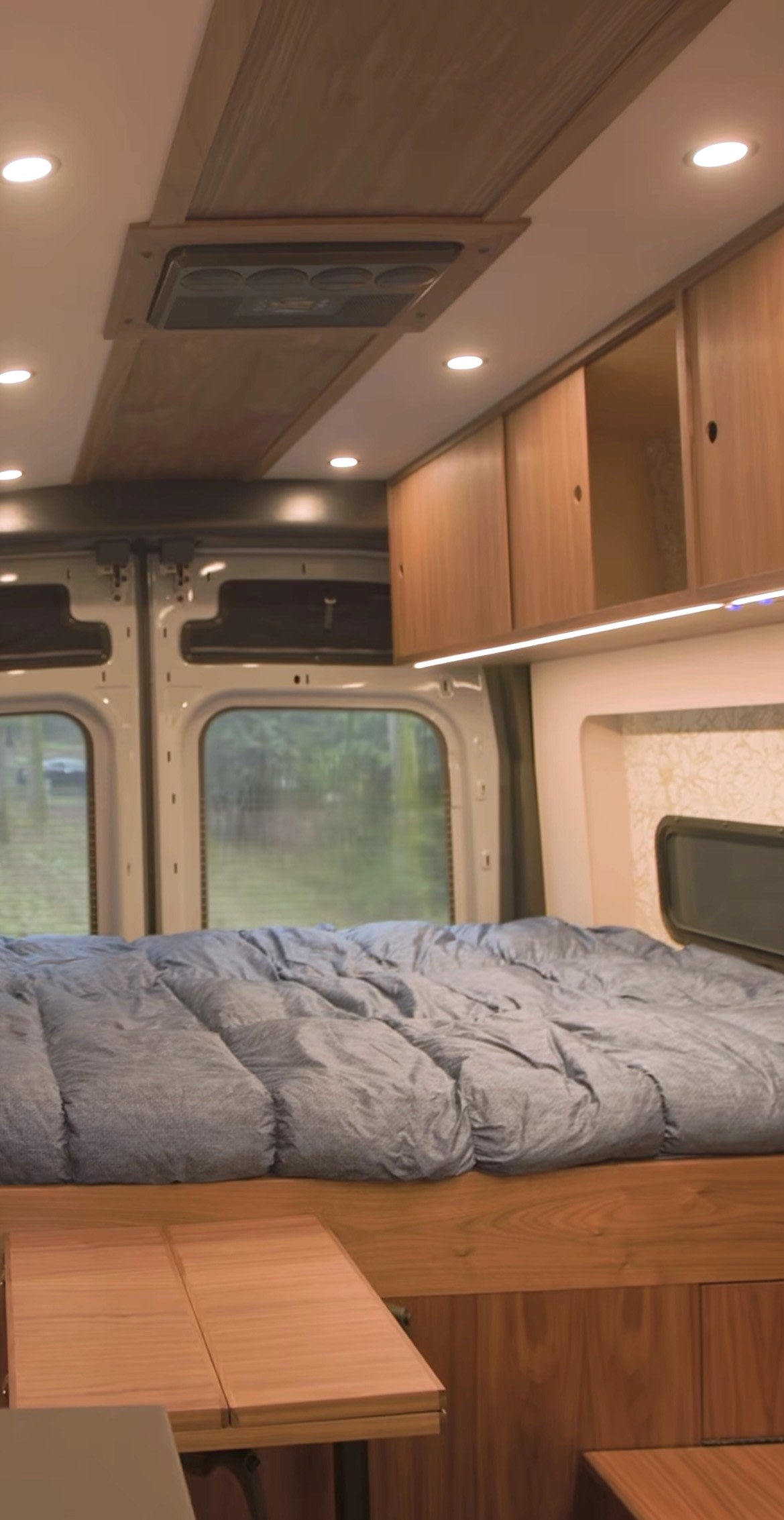 This RV rental interior features a cozy bed, wooden cabinetry, ceiling fan, and a foldable table, with windows offering nature views.