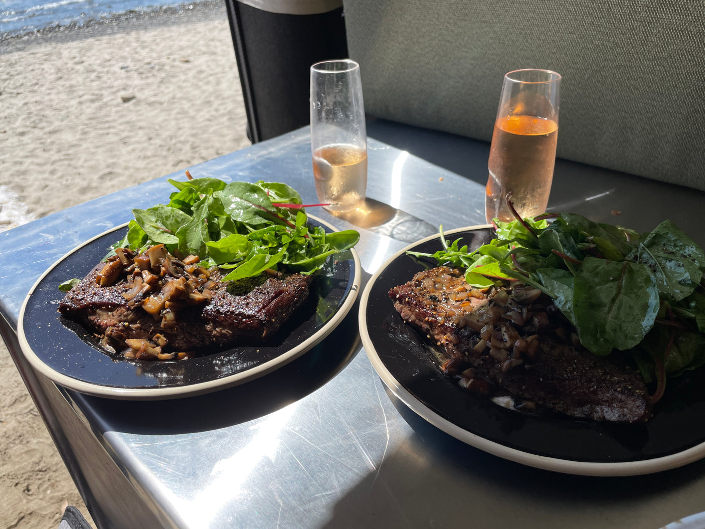 Gourmet beach dining with an RV rental: Two plates of steak & mushroom with salad, and wine, by the sea.
