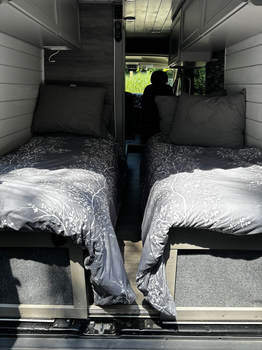 RV rental camper van interior with two comfortable single beds, grey patterned bedding, and narrow aisle leading to the front.