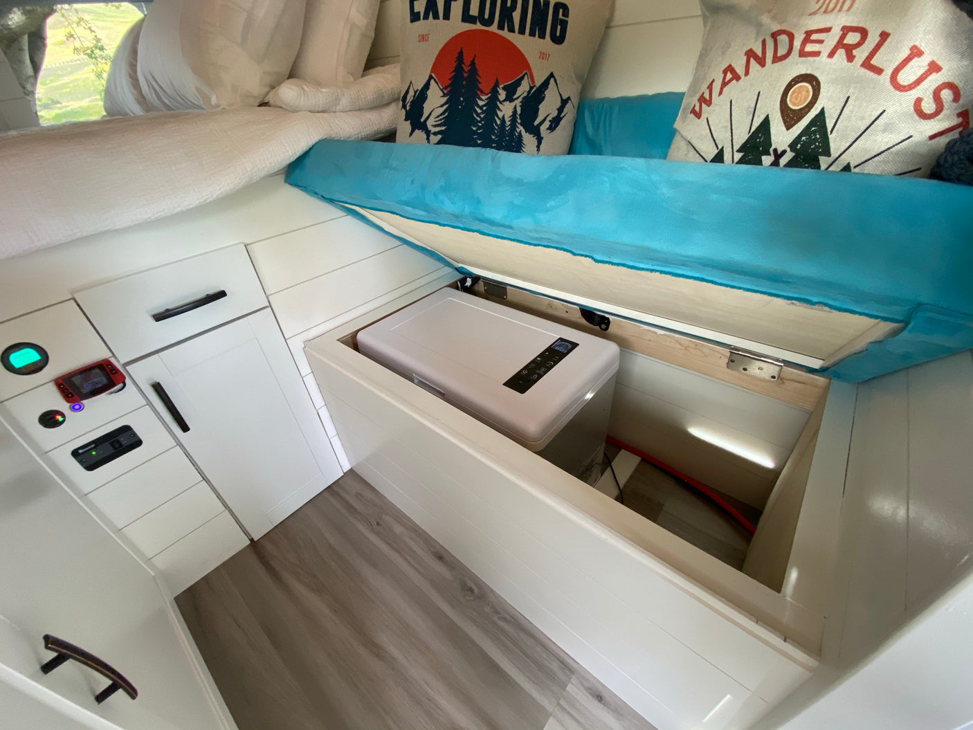 This RV rental features a cozy interior with a bed, under-bed storage revealing a portable fridge, and a functional control panel.