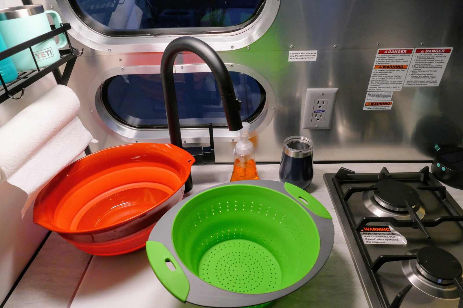 Inside an RV rental, showing a compact kitchen with a sink, gas stove, colorful collapsible bowls, and essential safety warnings.