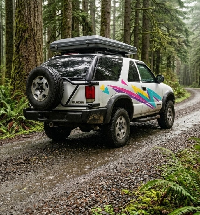 A white Chevy Blazer with a rooftop tent and vibrant decals on a forest trail. Ideal for your next RV rental adventure!|White adventure SUV with roof gear on a mountain path amidst wildflowers and a snowy peak. Ideal for an outdoor rv rental.|White SUV with roof-top tent on a scenic overlook at sunset
