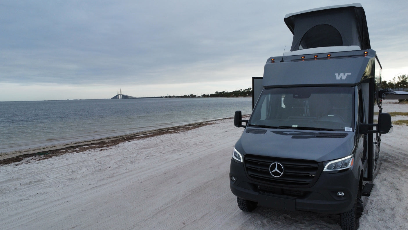 Grey pop-top RV rental on sandy beach by water with bridge view. Perfect for your next adventure!