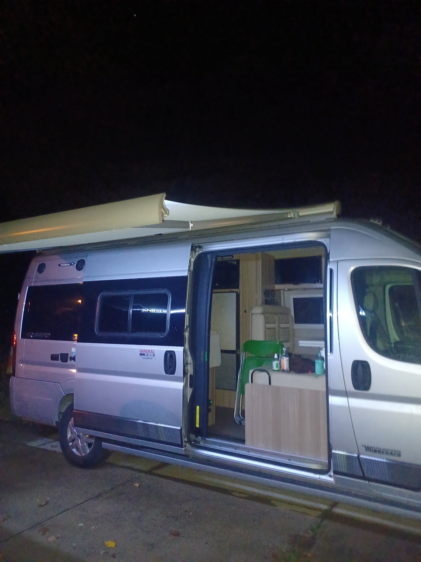 Night shot of a silver camper van, interior visible with a dining area & kitchen. An extended awning suggests camping. Great rv rental.