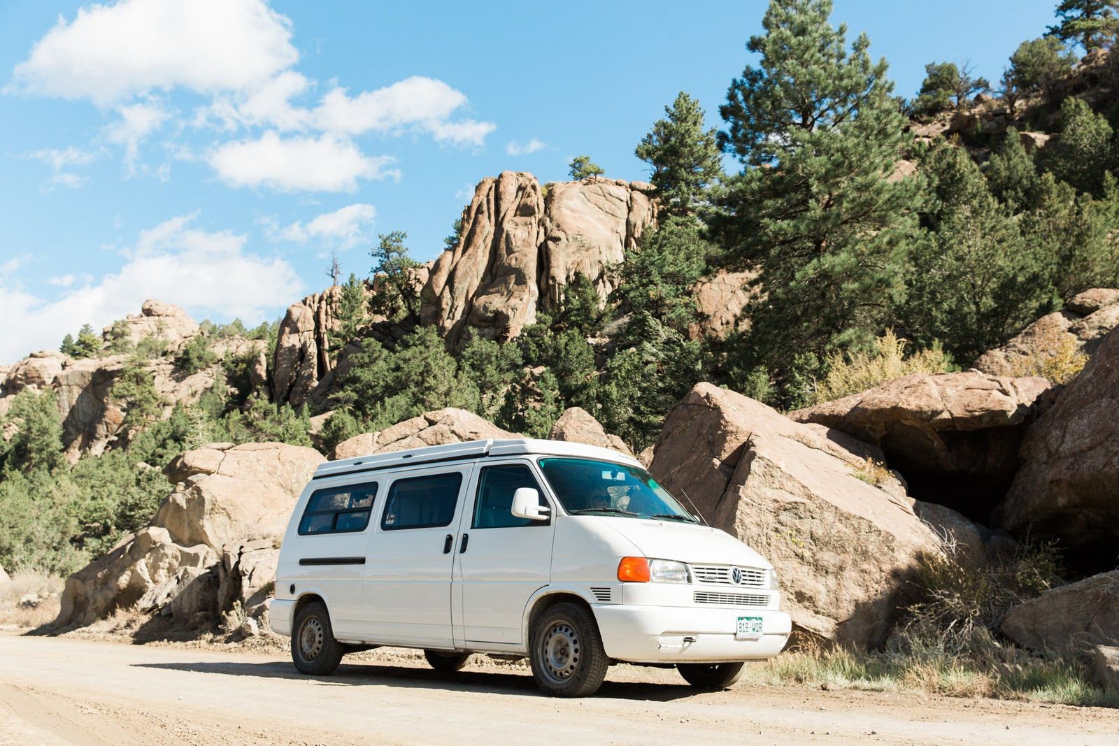 White VW camper van, like an RV rental, parked on a dirt road amidst scenic rock formations and pine trees under a blue sky.