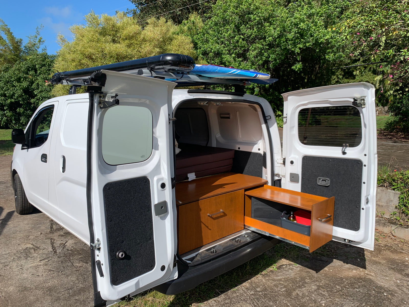 White camper van with side/rear doors open, revealing bed, cabinetry, and roof-mounted water tank/surfboards. Perfect for rv rental.
