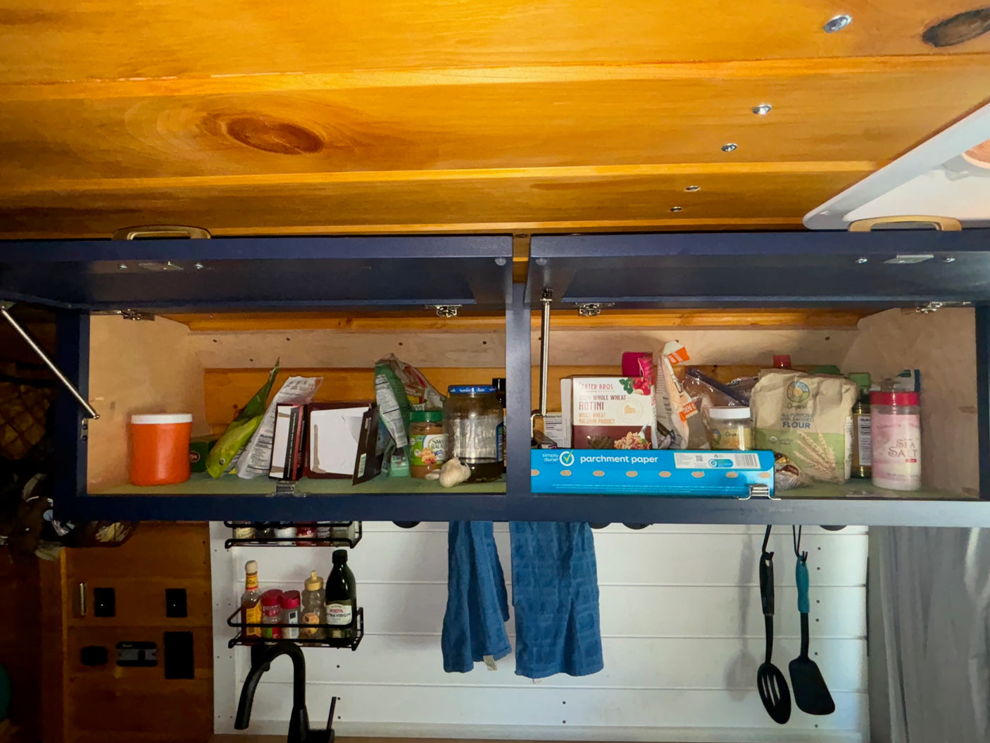 RV rental kitchen: open overhead cabinets reveal pantry essentials like food, parchment paper, spices, and utensils.