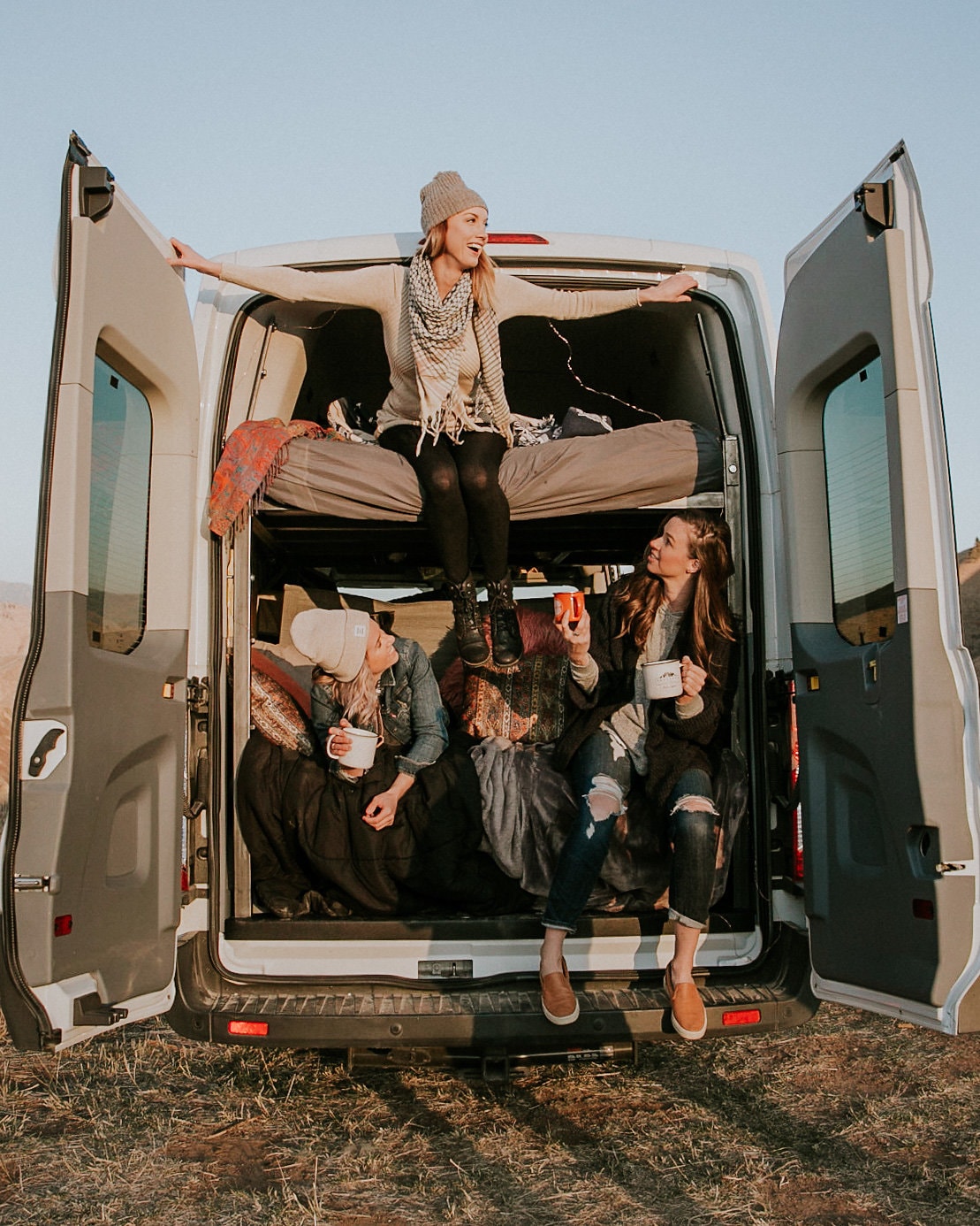 Friends enjoying an RV rental adventure. Three women relaxing in a converted van, sharing mugs, amidst nature.