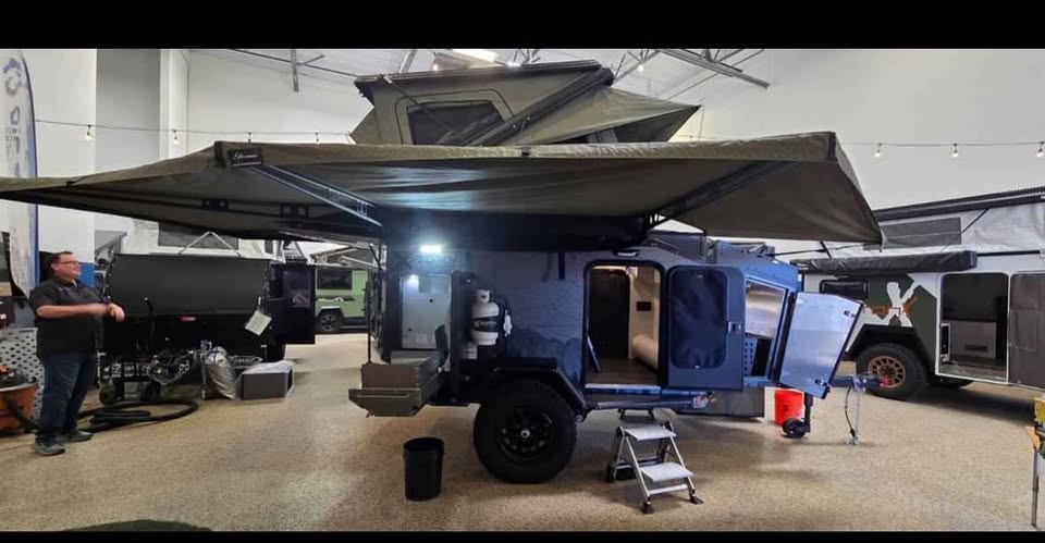 Driver-side view of the XPedition Voyager Sky XE indoors with the cabin door open, both 270 awnings extended overhead, rooftop tent up and a folding step stool at the doorway — photo 4