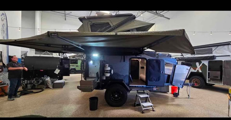 Driver-side view of the XPedition Voyager Sky XE indoors with the cabin door open, both 270 awnings extended overhead, rooftop tent up and a folding step stool at the doorway — photo 4