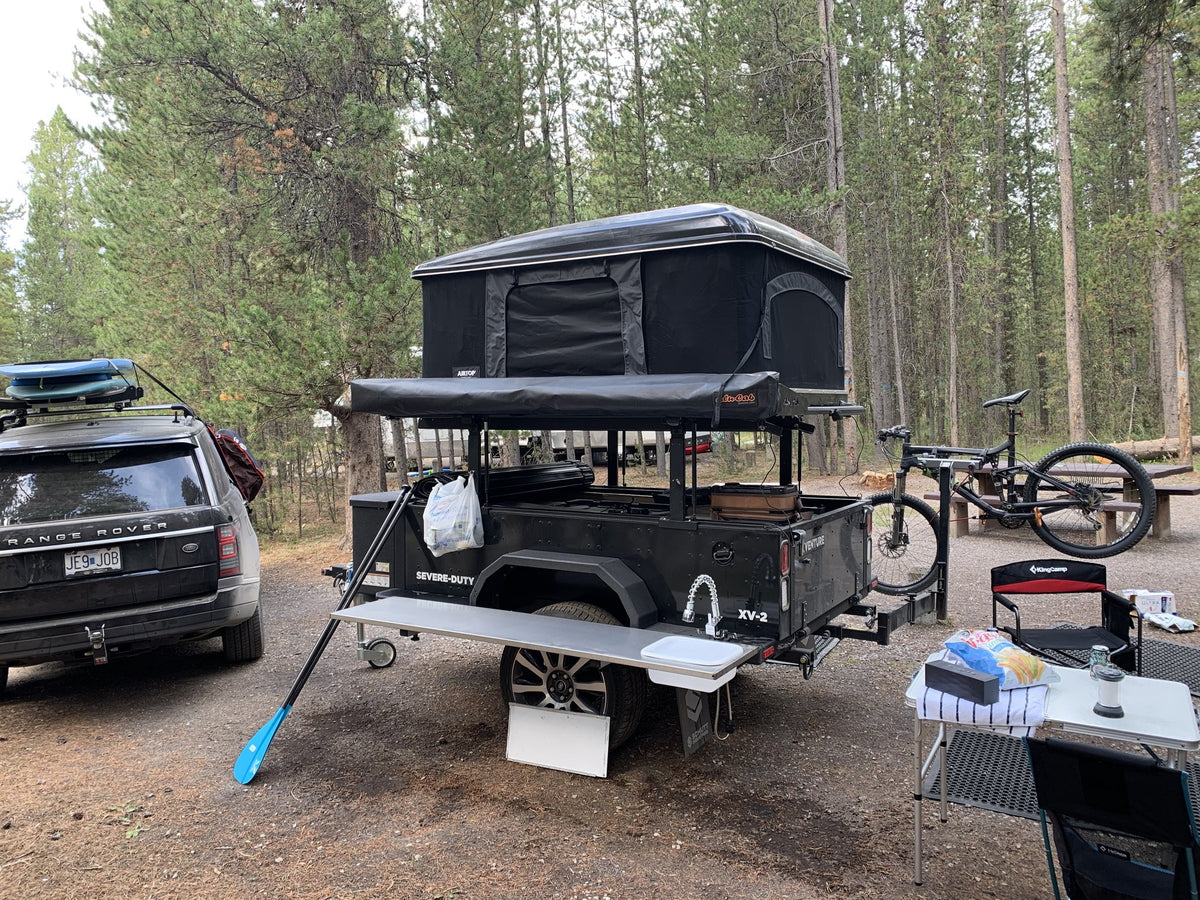 Trailer at a wooded campsite with the tent up, galley open and the service table set for cooking — photo 10