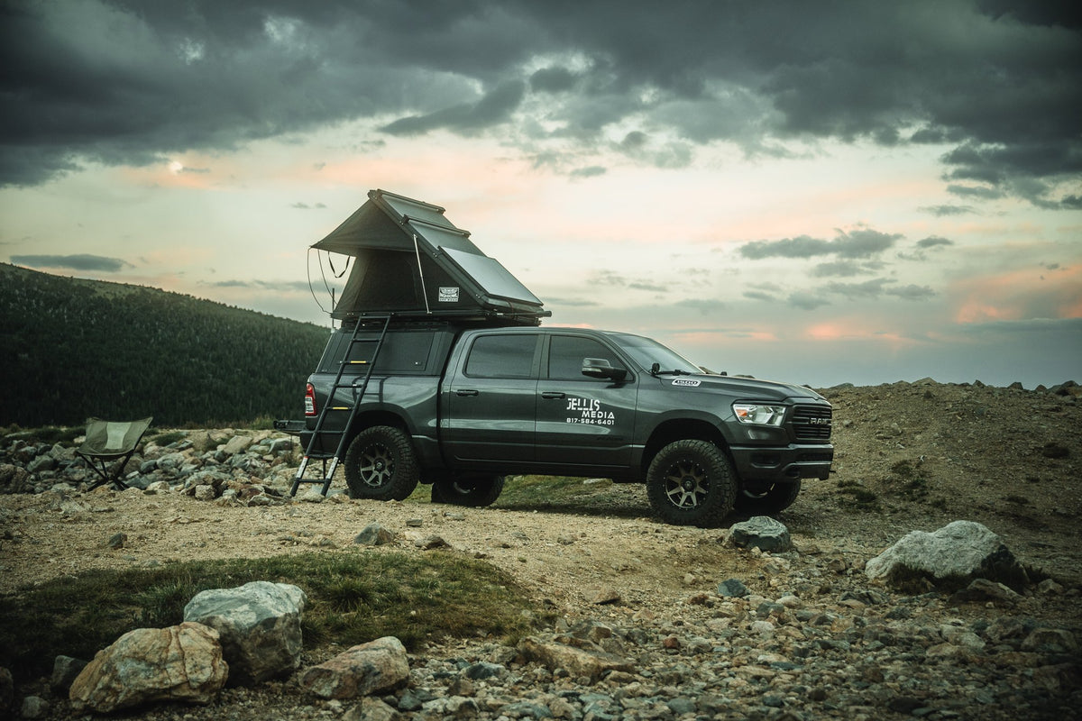 Passenger-side profile at a rocky mountain overlook at sunset, the rooftop tent open on top of the camper shell with its access ladder down and a camp chair set up alongside the truck — photo 2