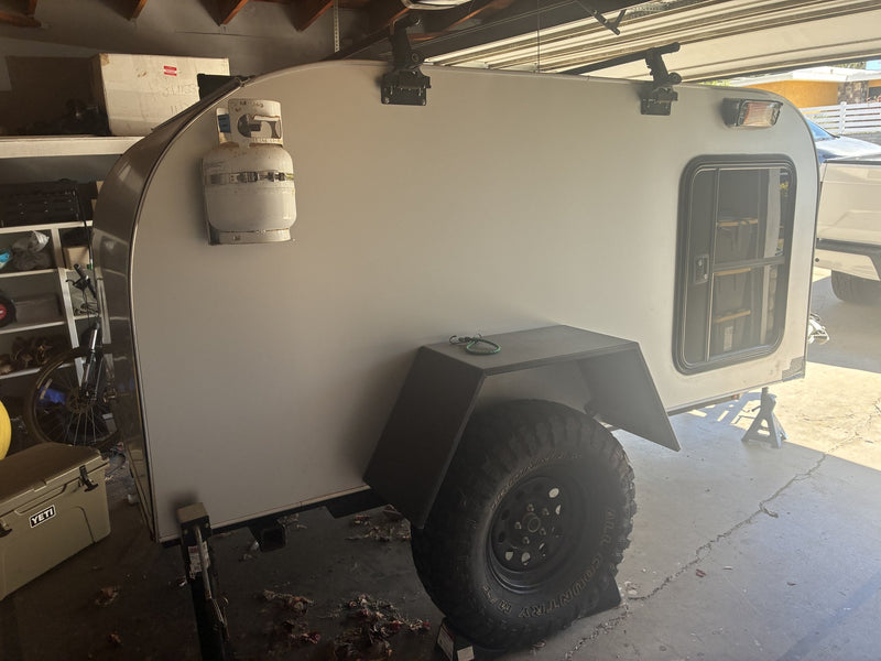 2020 Sierra Madre teardrop trailer, driver-side profile in a garage, showing the mounted propane tank, roof rack crossbars, black fender and all-terrain spare on a steel wheel — photo 1