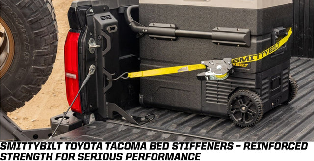 Smittybilt bed stiffener brace installed in a Toyota Tacoma truck bed, the black powder-coated internal brace bolted to the bed side ahead of the tailgate, with a portable fridge strapped down beside it and a spare tire visible over the bed rail