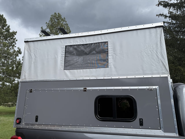 Side profile of the 2025 OVRLND Bivy pop-top camper mounted on a pickup bed with the roof raised, showing the white canvas pop-top with a mesh window, the grey aluminum side panel with a large flip-up gullwing hatch and a sliding window