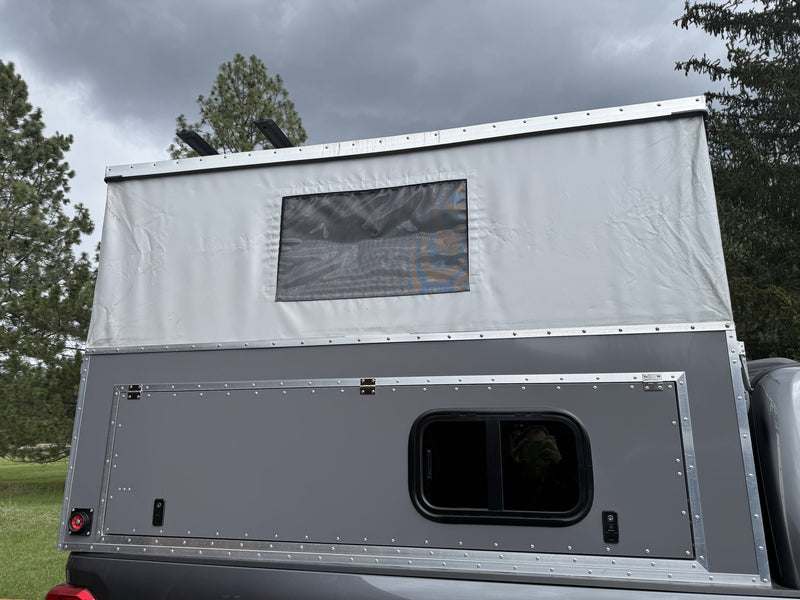 Side profile of the 2025 OVRLND Bivy pop-top camper mounted on a pickup bed with the roof raised, showing the white canvas pop-top with a mesh window, the grey aluminum side panel with a large flip-up gullwing hatch and a sliding window — photo 1
